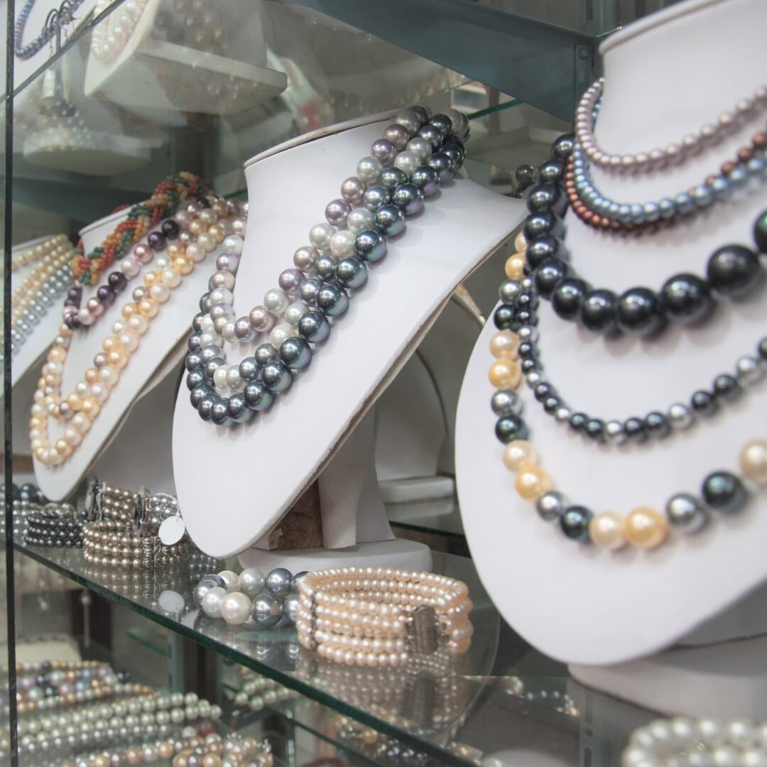 A display case with several white necklace stands showcasing multi-strand pearl necklaces in various colors and sizes, with matching pearl bracelets and jewelry pieces on the glass shelves below.
