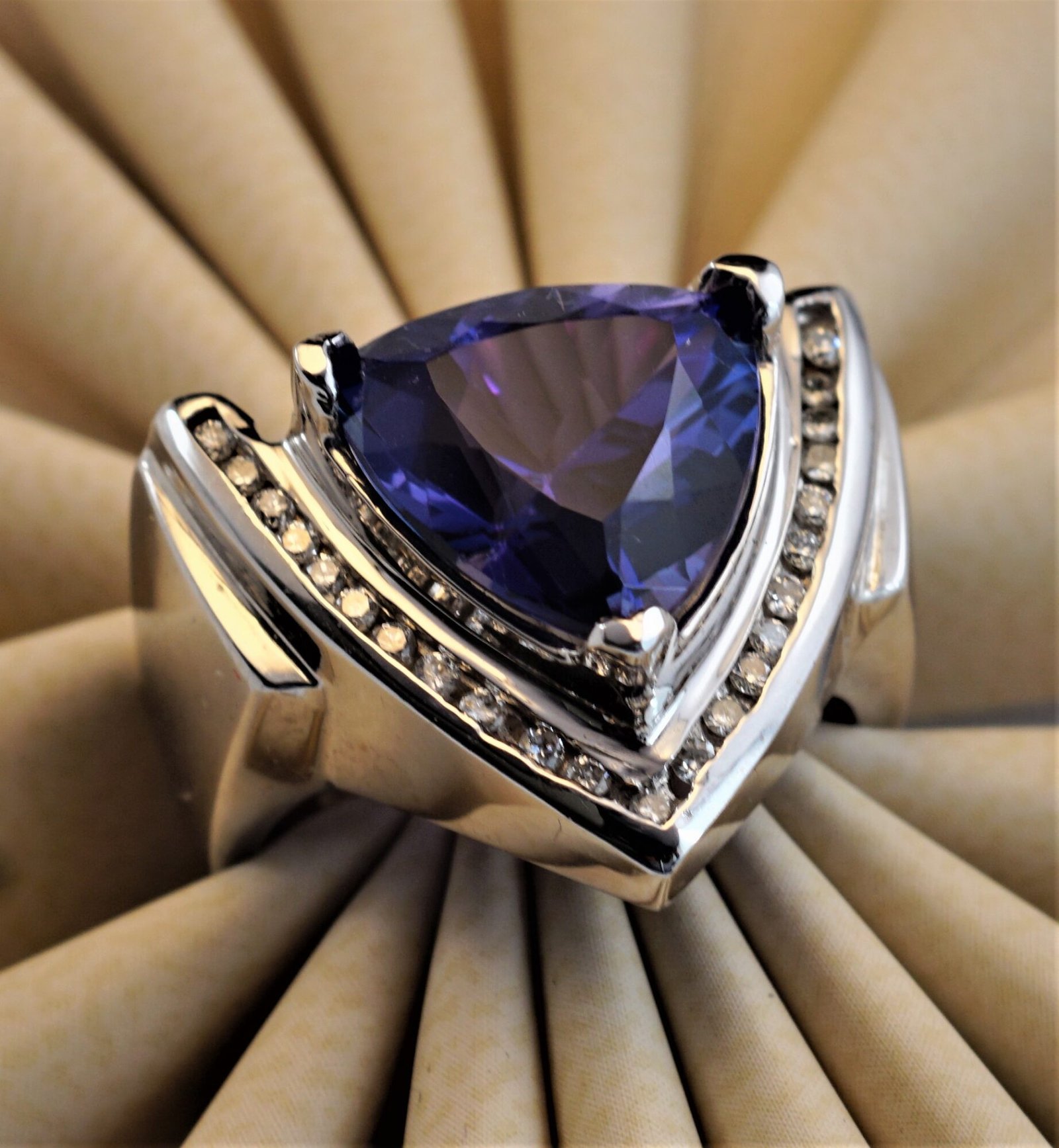 The Opulent Trillion-Cut Tanzanite & Diamond Ring in 14K White Gold features a stunning blue trillion-cut tanzanite flanked by diamonds, beautifully showcased on a beige pleated fabric background.