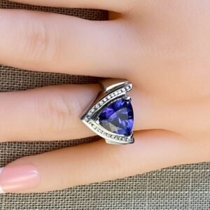 A hand displays the Opulent Trillion-Cut Tanzanite & Diamond Ring in 14K White Gold, featuring a large triangular purple tanzanite bordered by diamonds, resting on textured brown fabric.